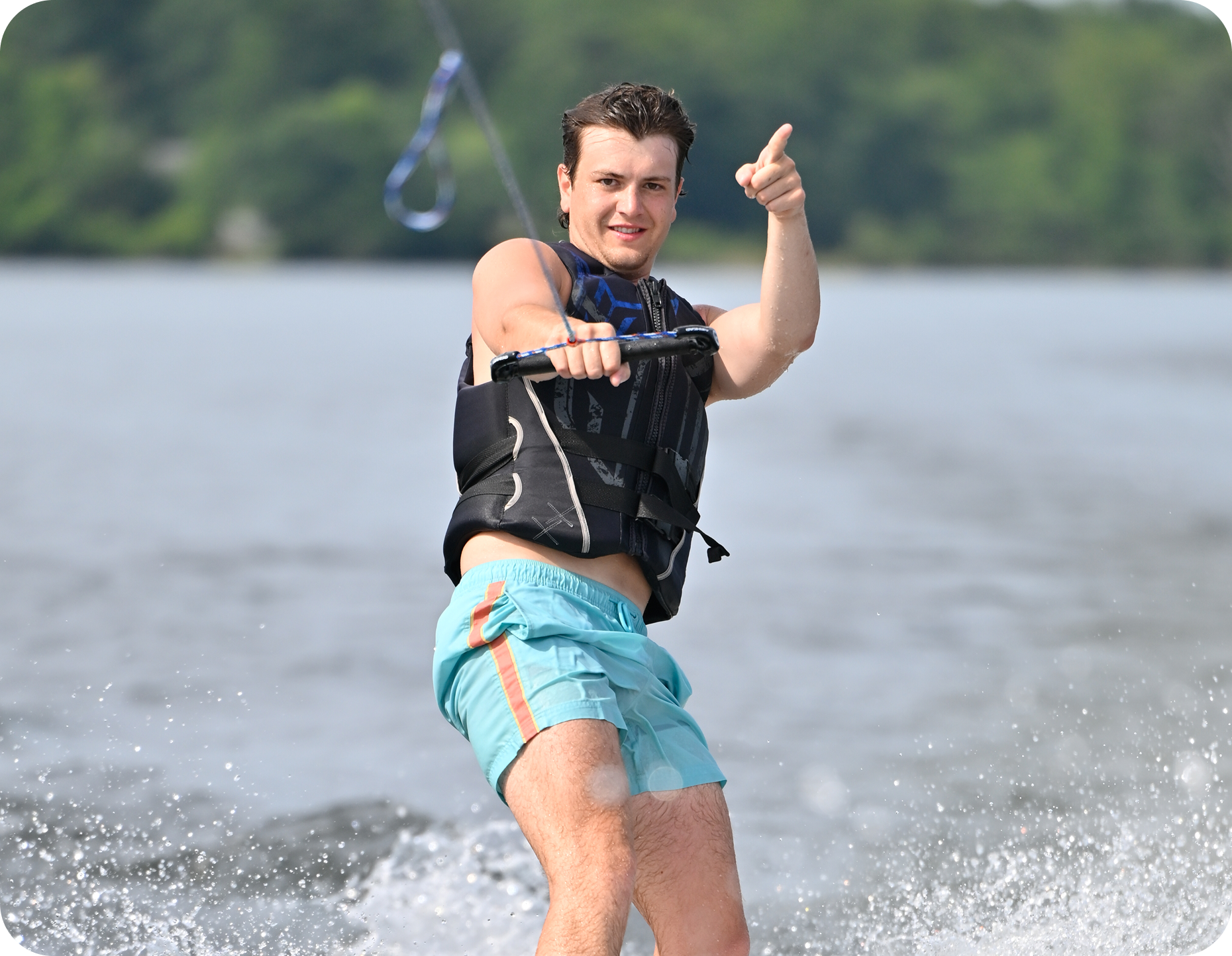A man in a life jacket and blue swim trunks is wakeboarding on a lake, holding a tow rope with one hand and pointing forward with the other. Water splashes around him and trees are visible in the background.